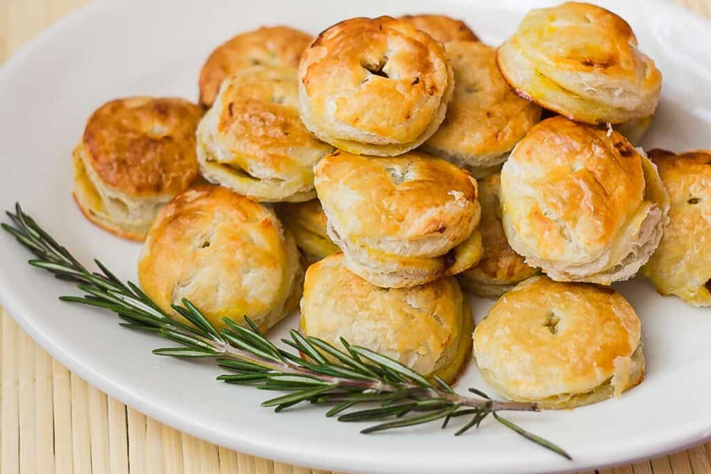 A plate of small, round, golden-brown puff pastries garnished with a sprig of rosemary.