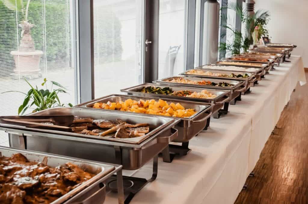 A buffet table with various dishes in chafing trays, including meats, vegetables, and pasta, is set up in a bright room with large windows and potted plants.