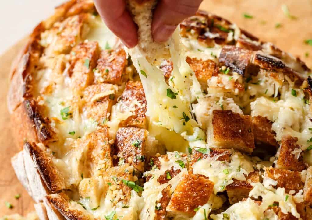 Close-up of a hand pulling melted cheese from a loaf of garlic bread topped with chopped herbs and browned cubes.