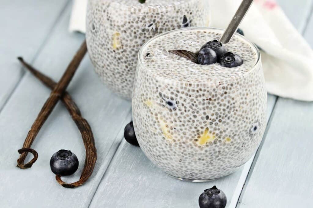 Two glasses of chia seed pudding topped with blueberries, placed on a wooden surface next to vanilla beans and scattered blueberries.