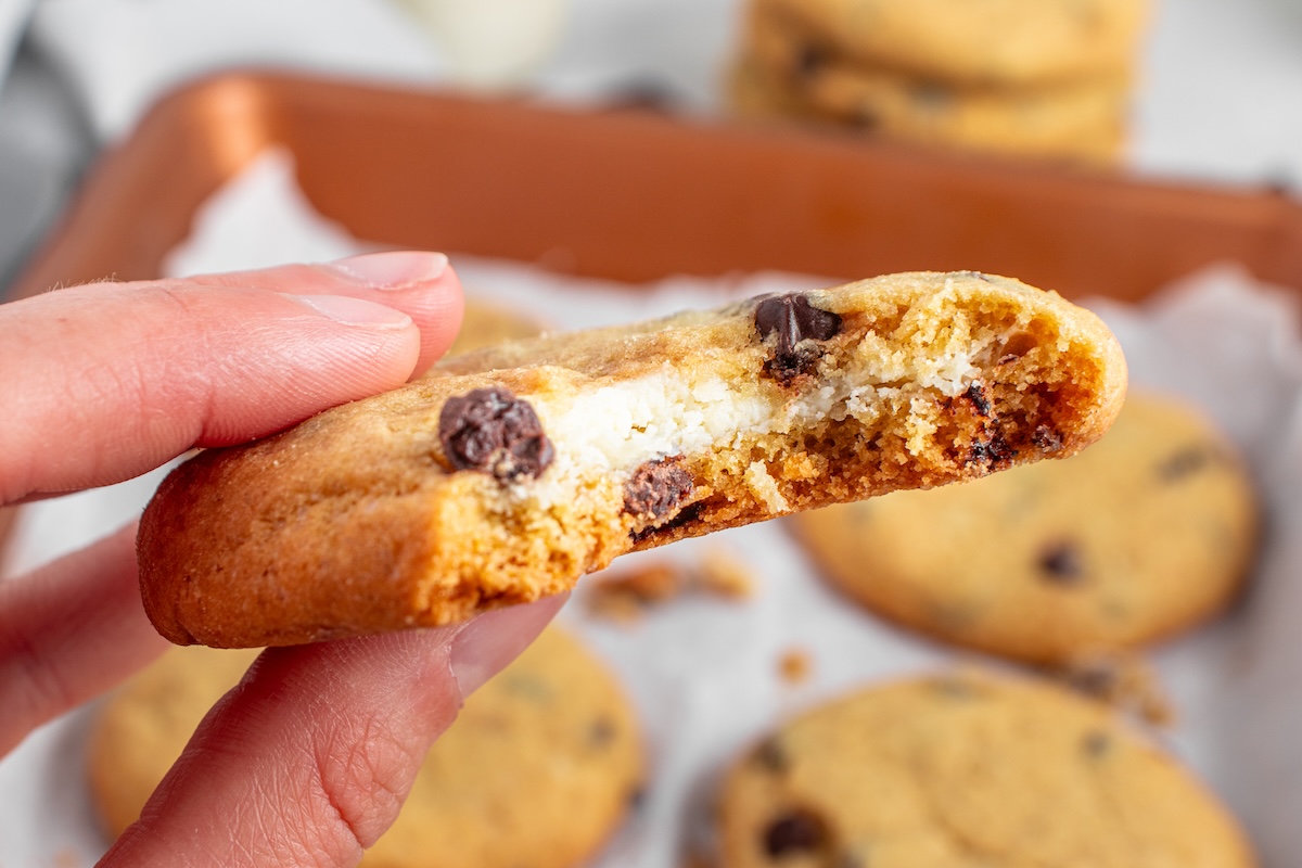 A close-up of a hand holding a chocolate chip cookie with a bite taken out, revealing a cheesecake filling inside. More cookies are visible in the background on a baking sheet.