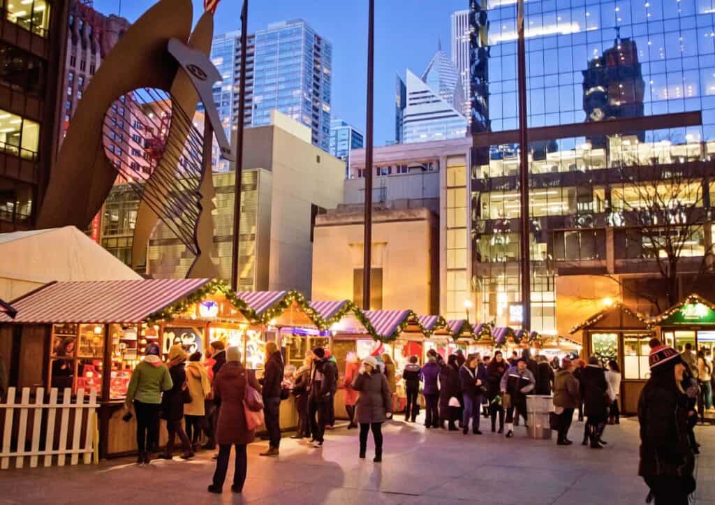 People browsing festive market stalls in a city square during the evening, with tall buildings in the background.