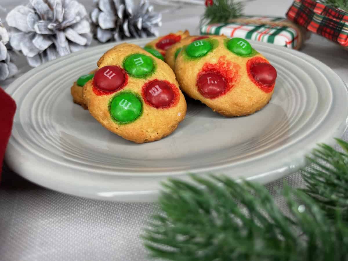 Two cookies topped with red and green chocolate candies on a gray plate, surrounded by pinecones and wrapped gifts.