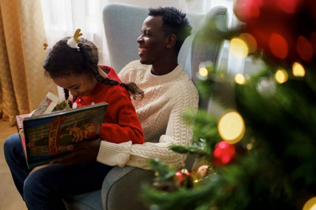 A man and a child sit in an armchair. The child is reading a book, and there's a decorated Christmas tree in the foreground.