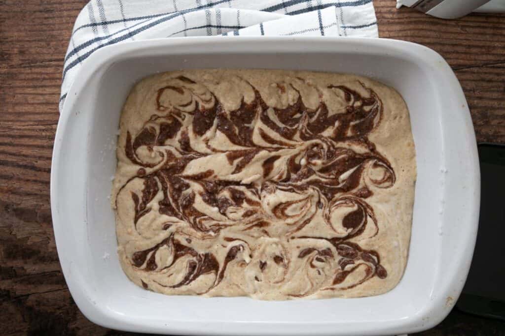 White baking dish with swirled chocolate and vanilla batter on a wooden surface, partially covered by a striped cloth.