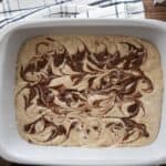 White baking dish with swirled chocolate and vanilla batter on a wooden surface, partially covered by a striped cloth.