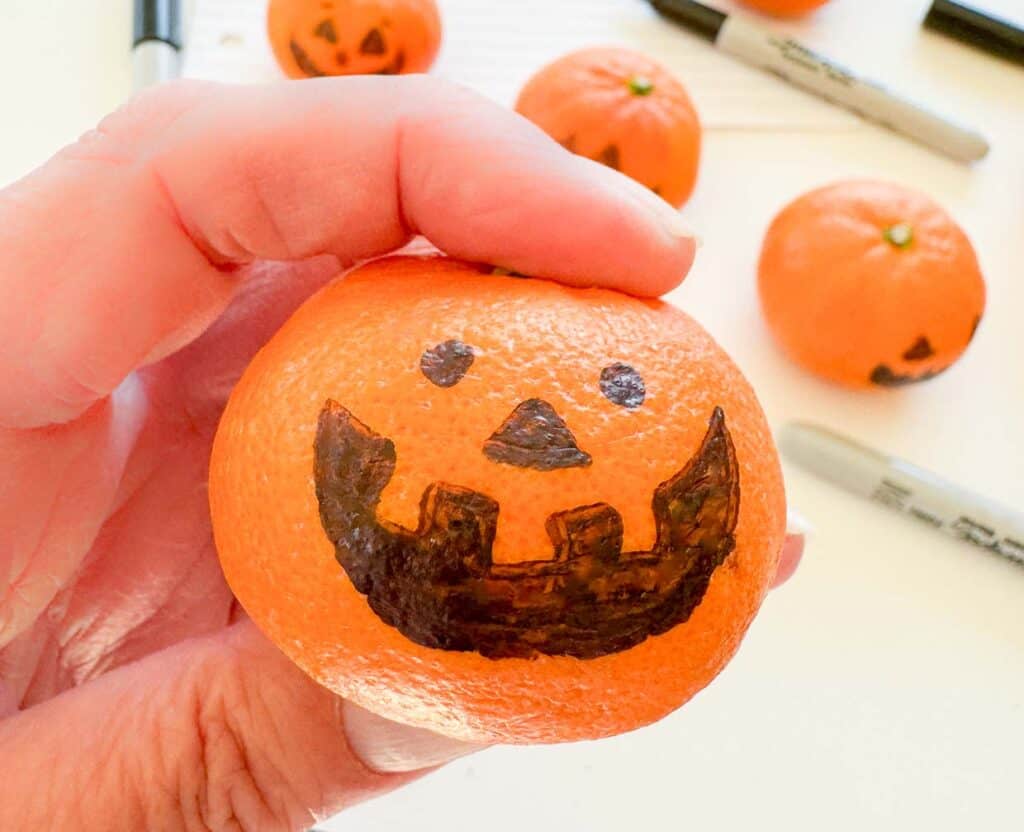 Hand holding a small orange with a jack-o'-lantern face drawn in black marker. Other decorated oranges and markers are in the background.