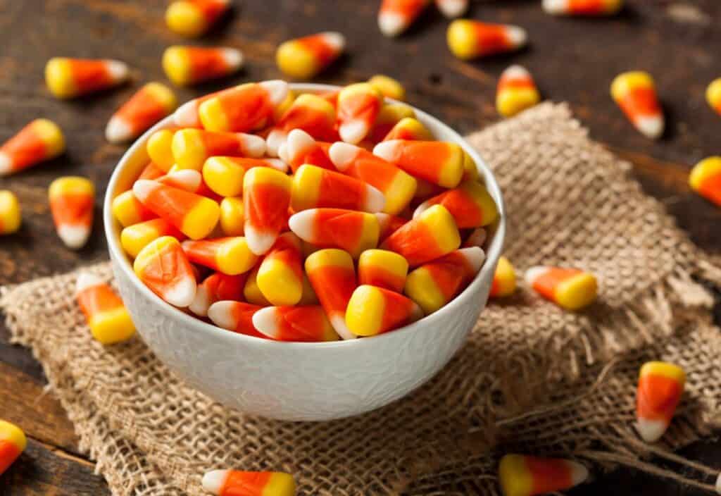 A white bowl filled with candy corn pieces sits on a piece of burlap, surrounded by more candy corn on a wooden surface.