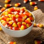 A white bowl filled with candy corn on a piece of burlap, surrounded by pieces of candy corn on a wooden surface.