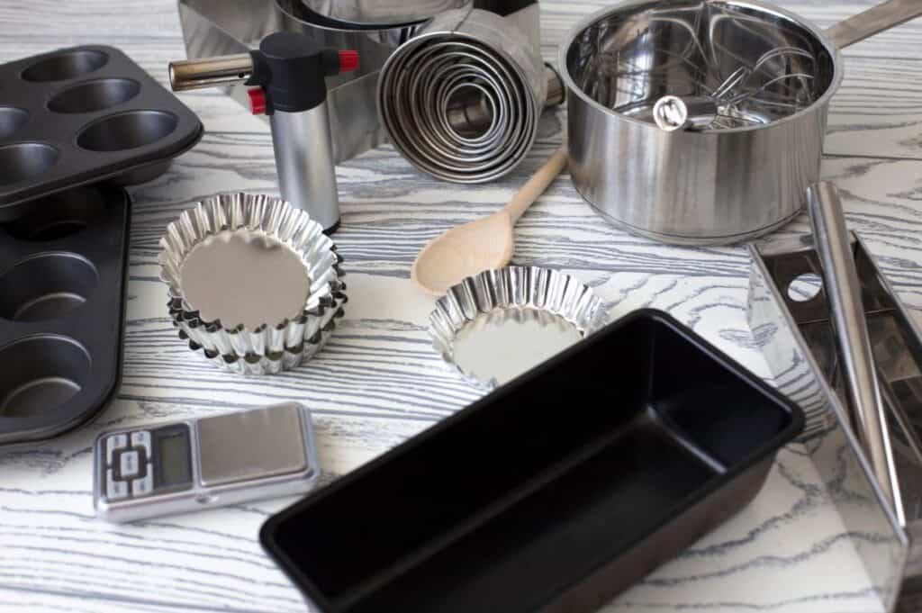 Assorted baking tools include muffin and loaf pans, measuring spoons, tart molds, a saucepan, a wooden spoon, a kitchen torch, and a digital scale, arranged on a gray patterned surface.