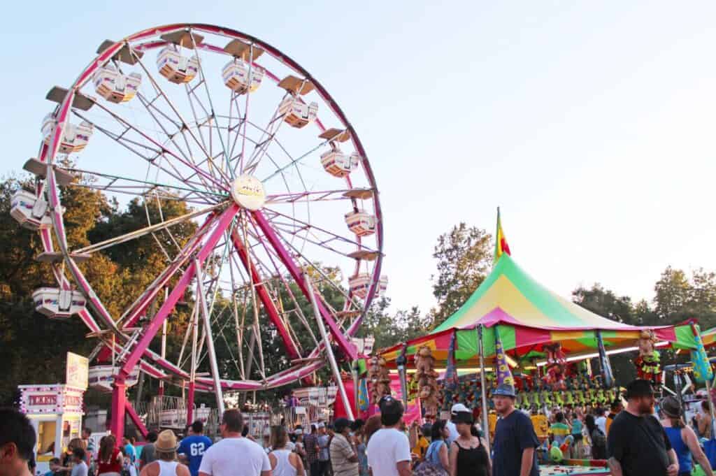 A busy fairground scene with a large Ferris wheel on the left and a colorful tent with games on the right. People are walking around enjoying the fair. Trees are visible in the background.