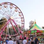 A bustling fair with a Ferris wheel, colorful tent, and people enjoying the attractions under a clear sky.