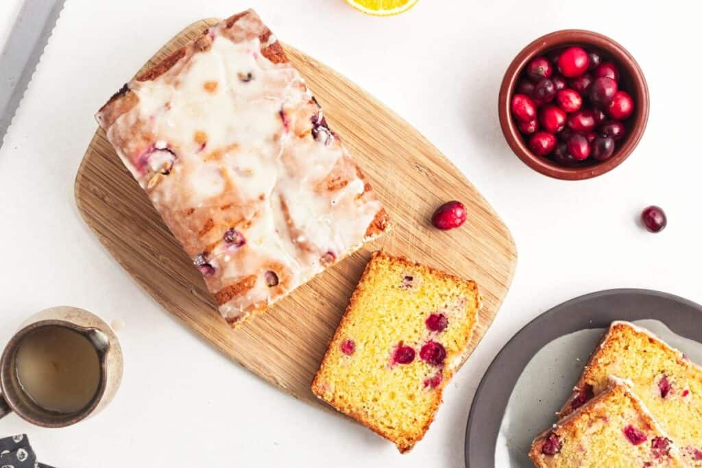 Iced cranberry loaf on a wooden board, surrounded by a bowl of cranberries, a cup of coffee, and a plate with slices on a white surface.