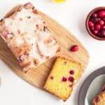 Iced cranberry loaf on a wooden board, surrounded by a bowl of cranberries, a cup of coffee, and a plate with slices on a white surface.