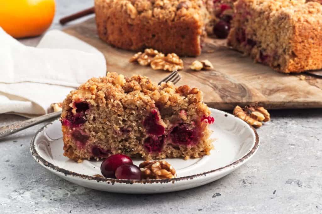 A slice of cranberry walnut crumb cake on a plate with additional cranberries and walnuts. The rest of the cake is on a wooden board in the background.