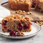 A slice of cranberry walnut crumb cake on a plate with additional cranberries and walnuts. The rest of the cake is on a wooden board in the background.