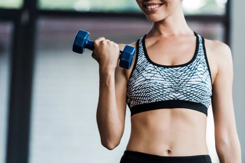 Person in a patterned sports bra lifting a blue dumbbell indoors, torso visible.