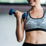 Person in a patterned sports bra lifting a blue dumbbell indoors, torso visible.