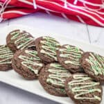 A plate of chocolate cookies topped with white icing and green sprinkles. A red and white striped cloth is in the background.