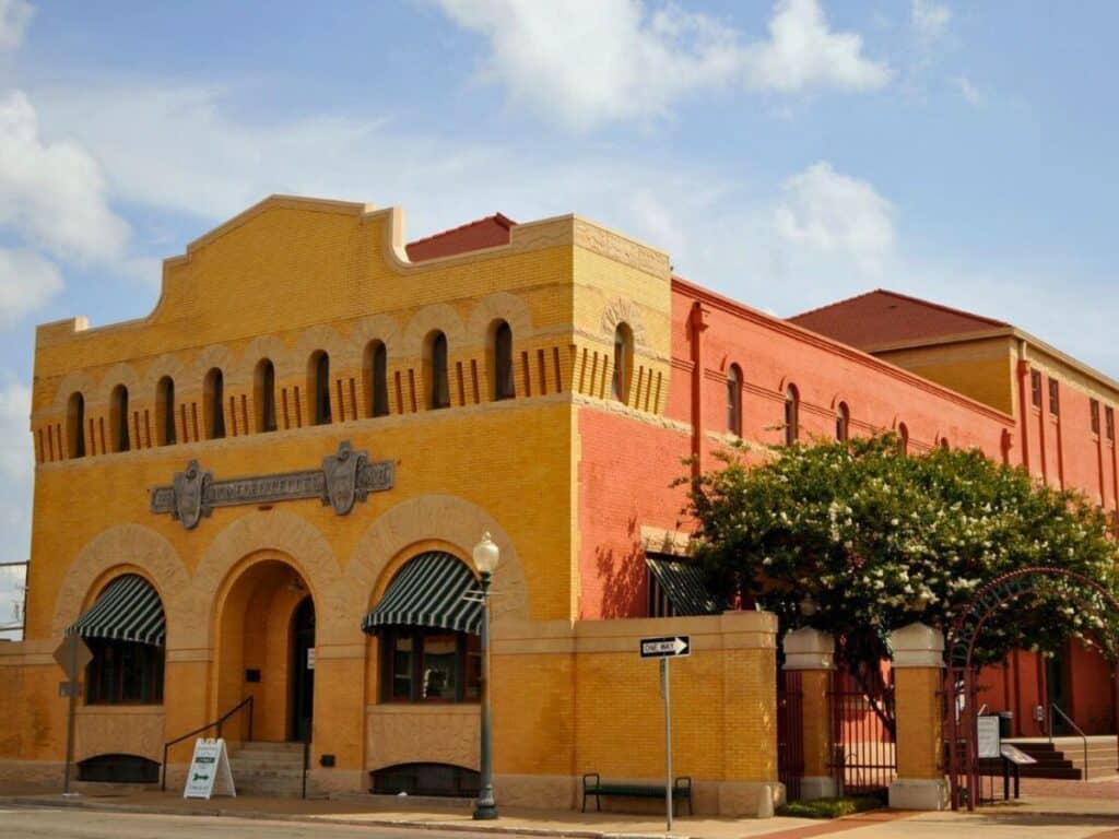 A historic red and yellow brick building with arched windows and a sign above the entrance, this iconic spot in Waco is surrounded by trees and street signs, offering a glimpse into the city's rich heritage among its many attractions.