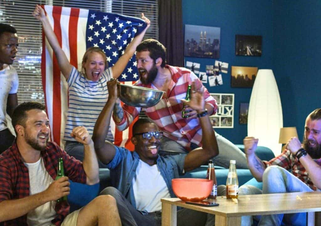 A group of people celebrate enthusiastically with drinks and snacks in a room decorated with an American flag and photos.