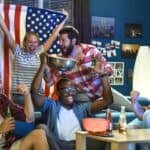 A group of people celebrate enthusiastically with drinks and snacks in a room decorated with an American flag and photos.