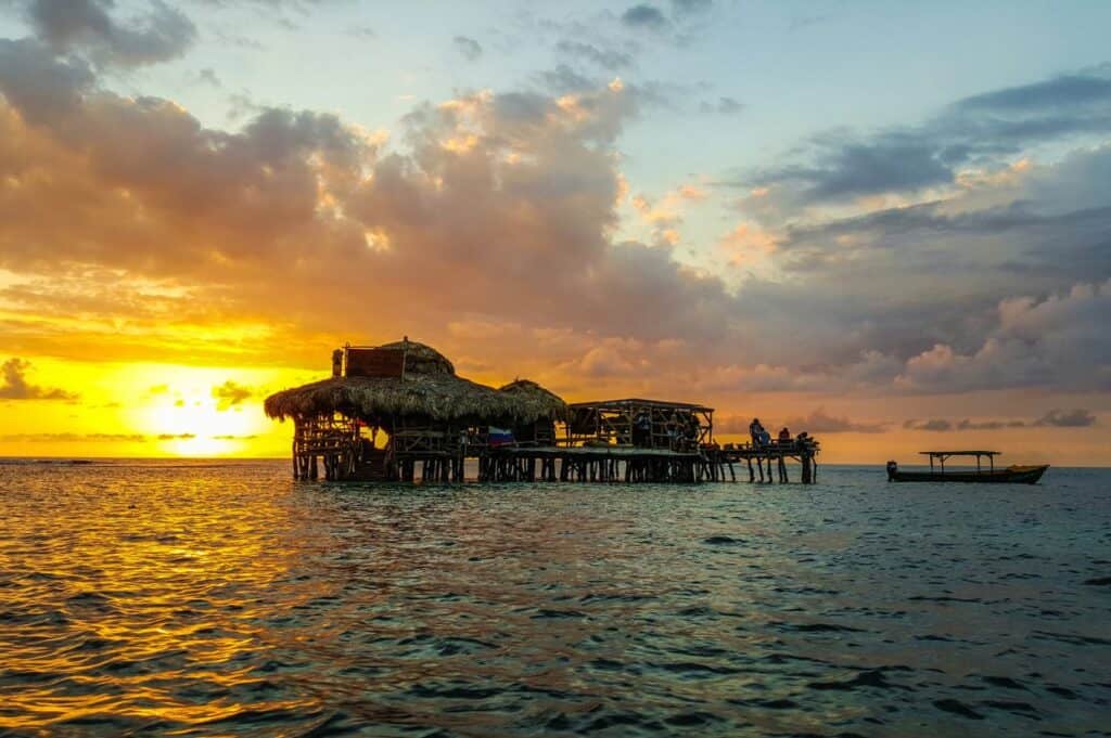 A wooden structure on stilts with a thatched roof stands over water at sunset, echoing the charm of the best beach bars in the Caribbean, with a small boat nearby under a partly cloudy sky.