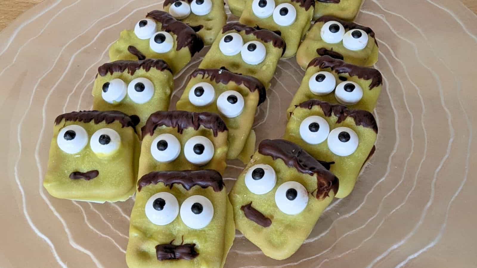 Plate of rectangular cookies decorated with green frosting, googly eyes, and chocolate hair resembling cartoon zombie faces.