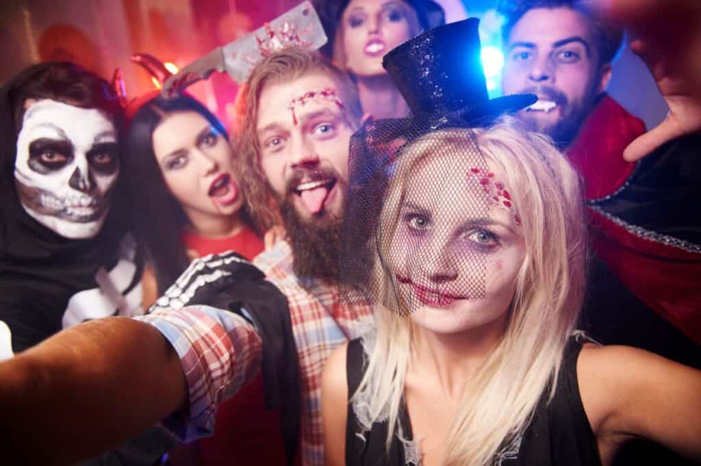 A group of people in Halloween costumes, including a skeleton, zombie, and vampire, posing for a selfie at a party with colorful lights in the background.