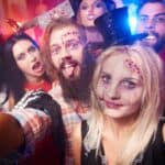 A group of people in Halloween costumes, including a skeleton, zombie, and vampire, posing for a selfie at a party with colorful lights in the background.