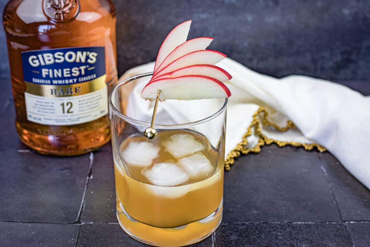 A glass of Gibson’s Apple Pie Old Fashioned garnished with sliced apples on a skewer.