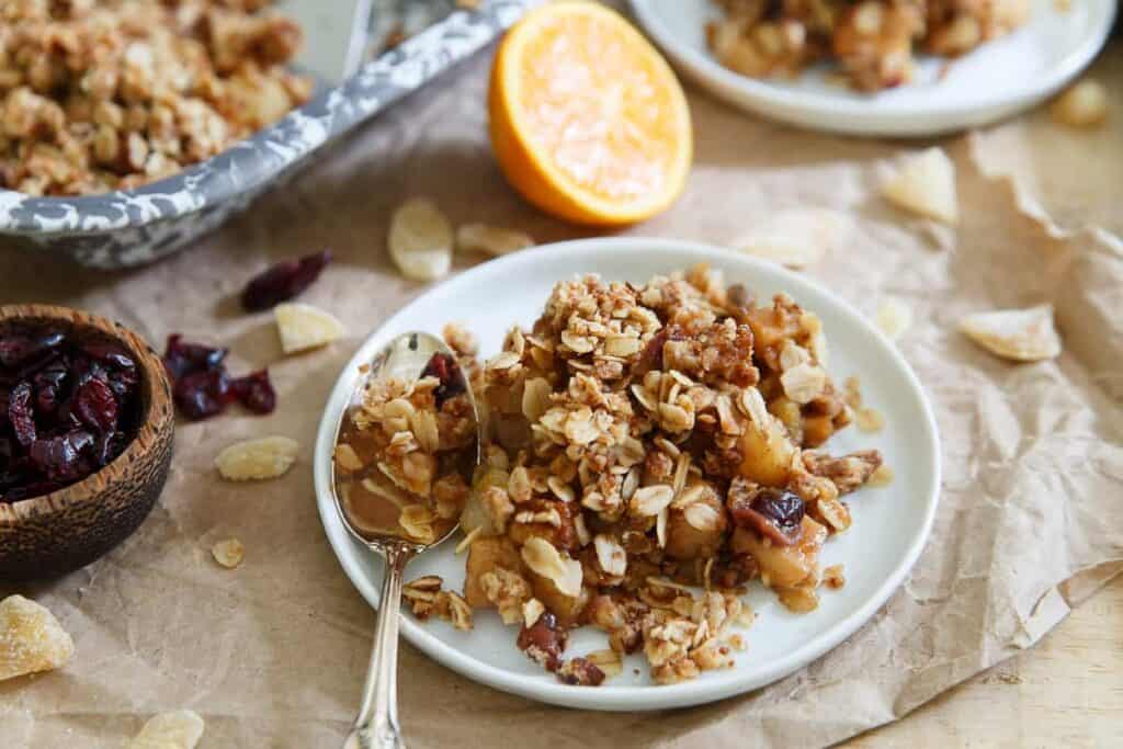 A plate of granola with nuts and dried fruit on a table, next to a bowl of dried cranberries and an orange half.