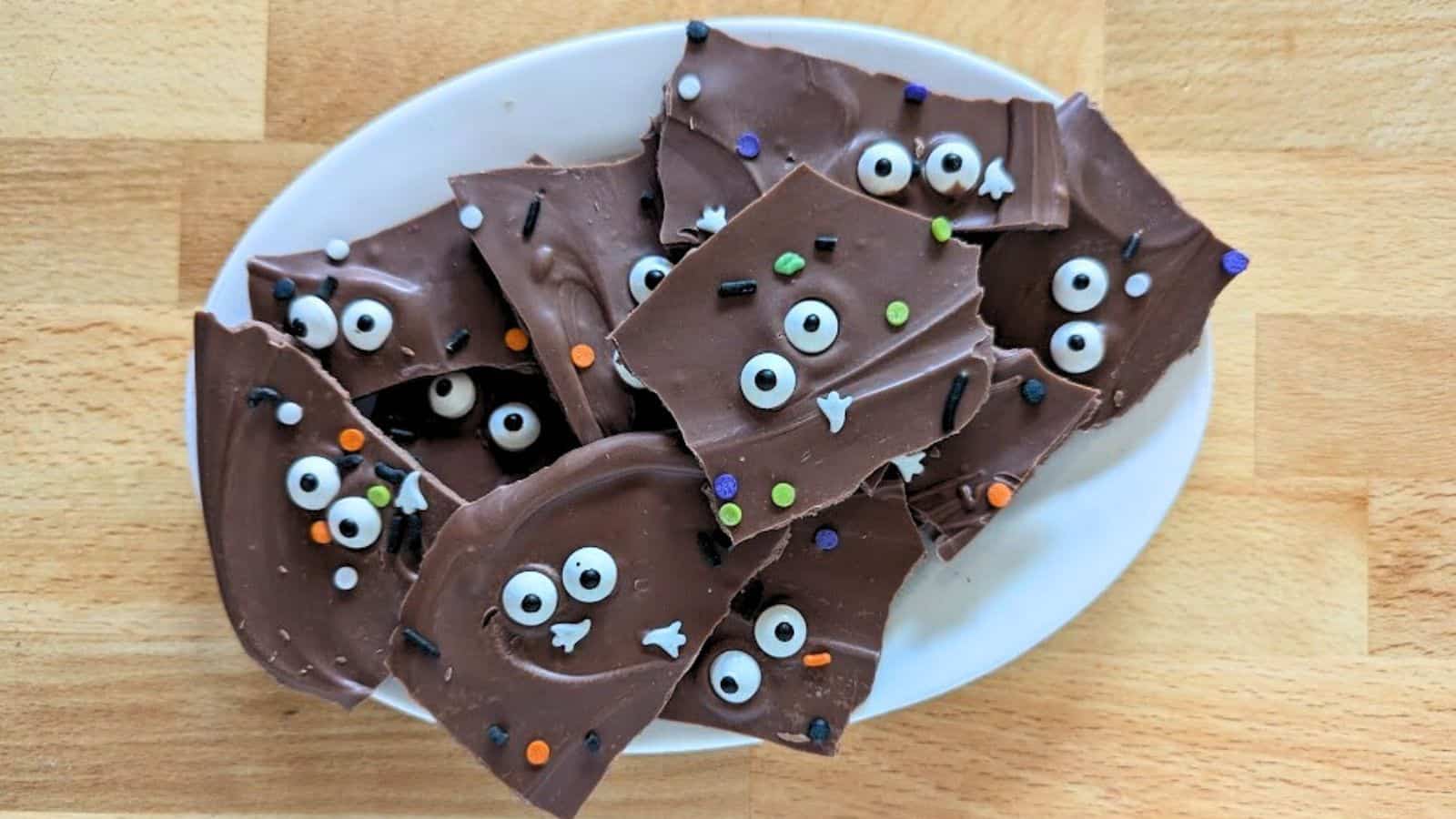 A white plate holds pieces of chocolate bark topped with candy eyes and colorful sprinkles, placed on a wooden surface.