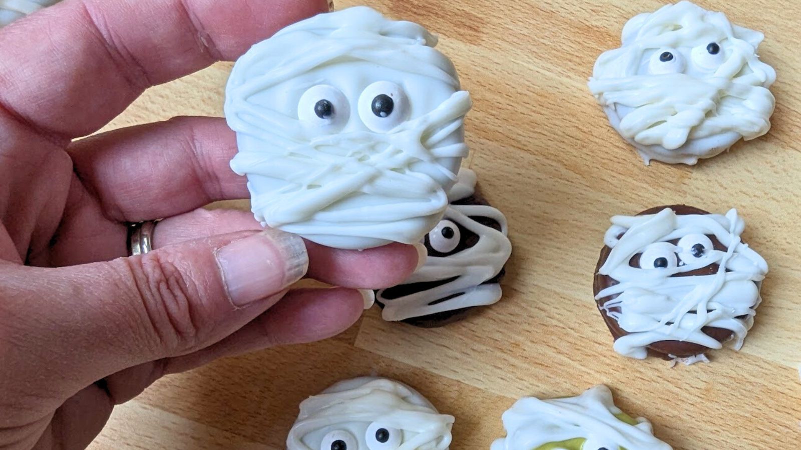 A hand holding a cookie decorated with white icing to resemble a mummy. Similar cookies are on the wooden surface below.