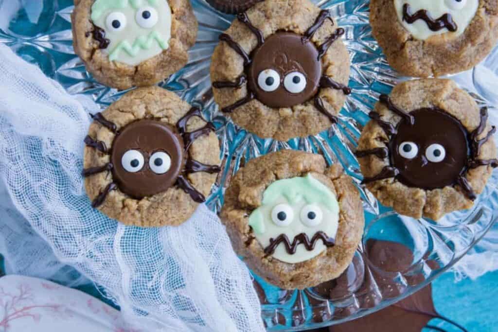 Cookies decorated with cartoonish monster and spider faces, featuring chocolate and icing details, arranged on a glass plate.