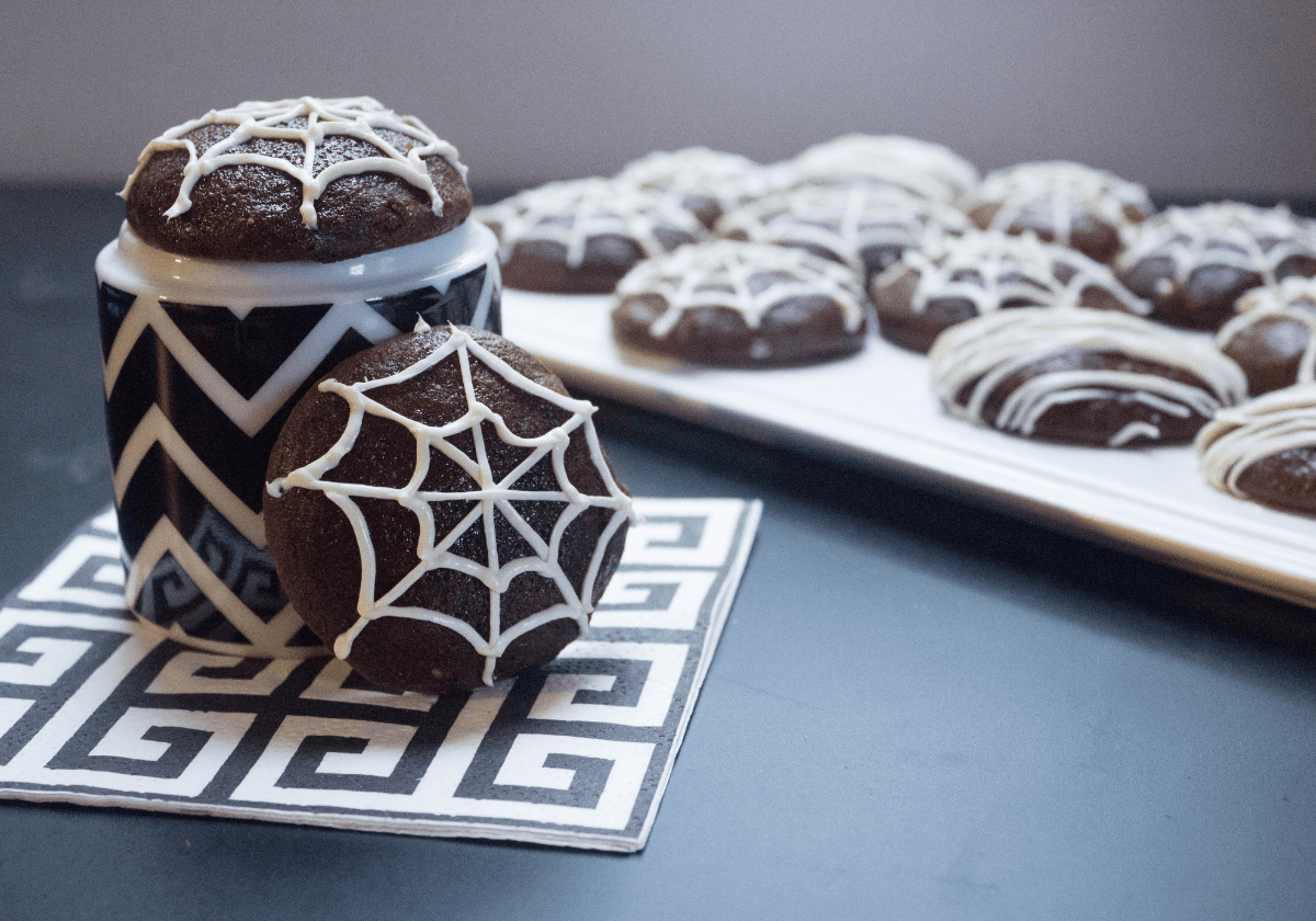 Chocolate cookies with icing on top decorated to look like spiderwebs, sitting in front of a black and white cookie jar.