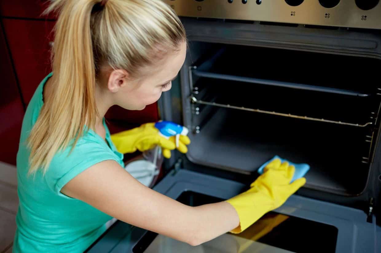 A person wearing yellow gloves is cleaning an open oven with a spray bottle and cloth.