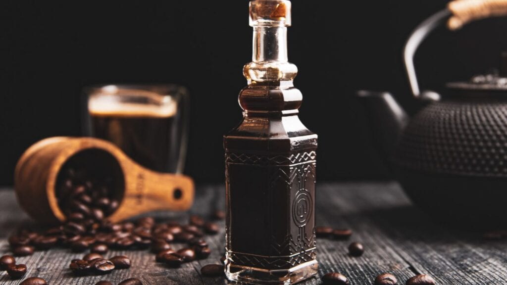 A bottle of dark liquid on a wooden table surrounded by coffee beans, with a coffee cup and teapot in the background.