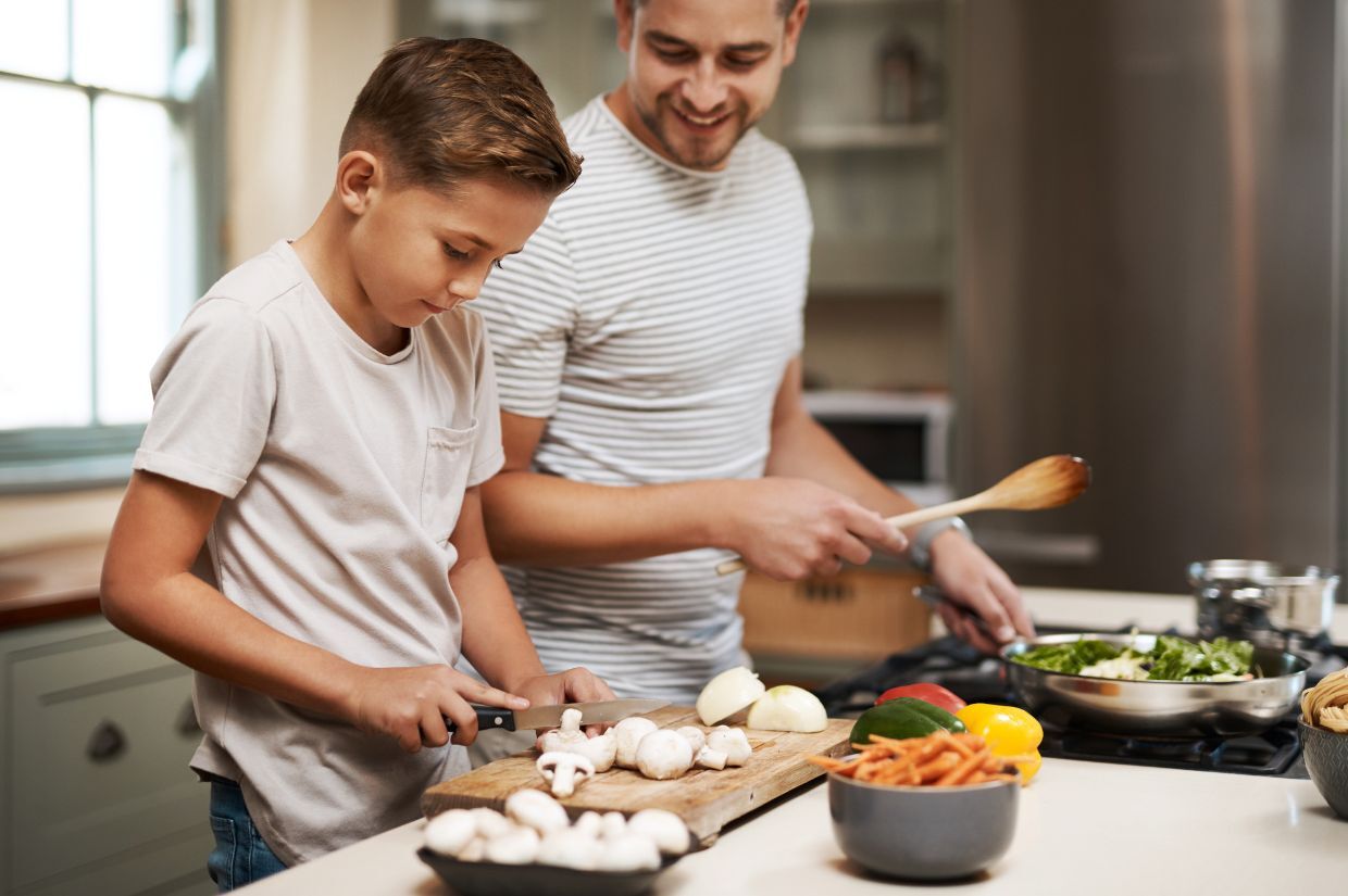 A man and a boy are preparing food in a kitchen. The boy is chopping mushrooms while the man stirs vegetables in a pan. Various ingredients are on the counter.