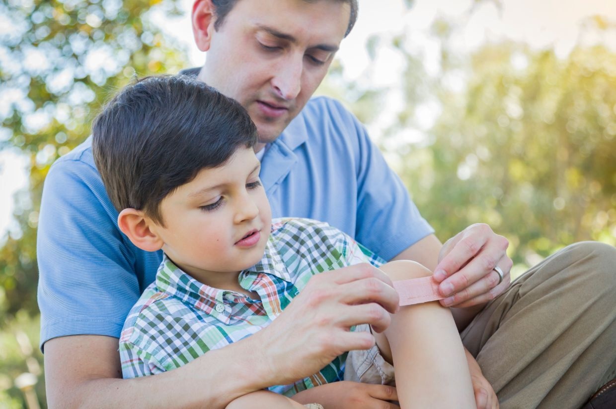 A man in a light blue shirt bandages a young boy's knee outdoors. The boy is wearing a plaid shirt.