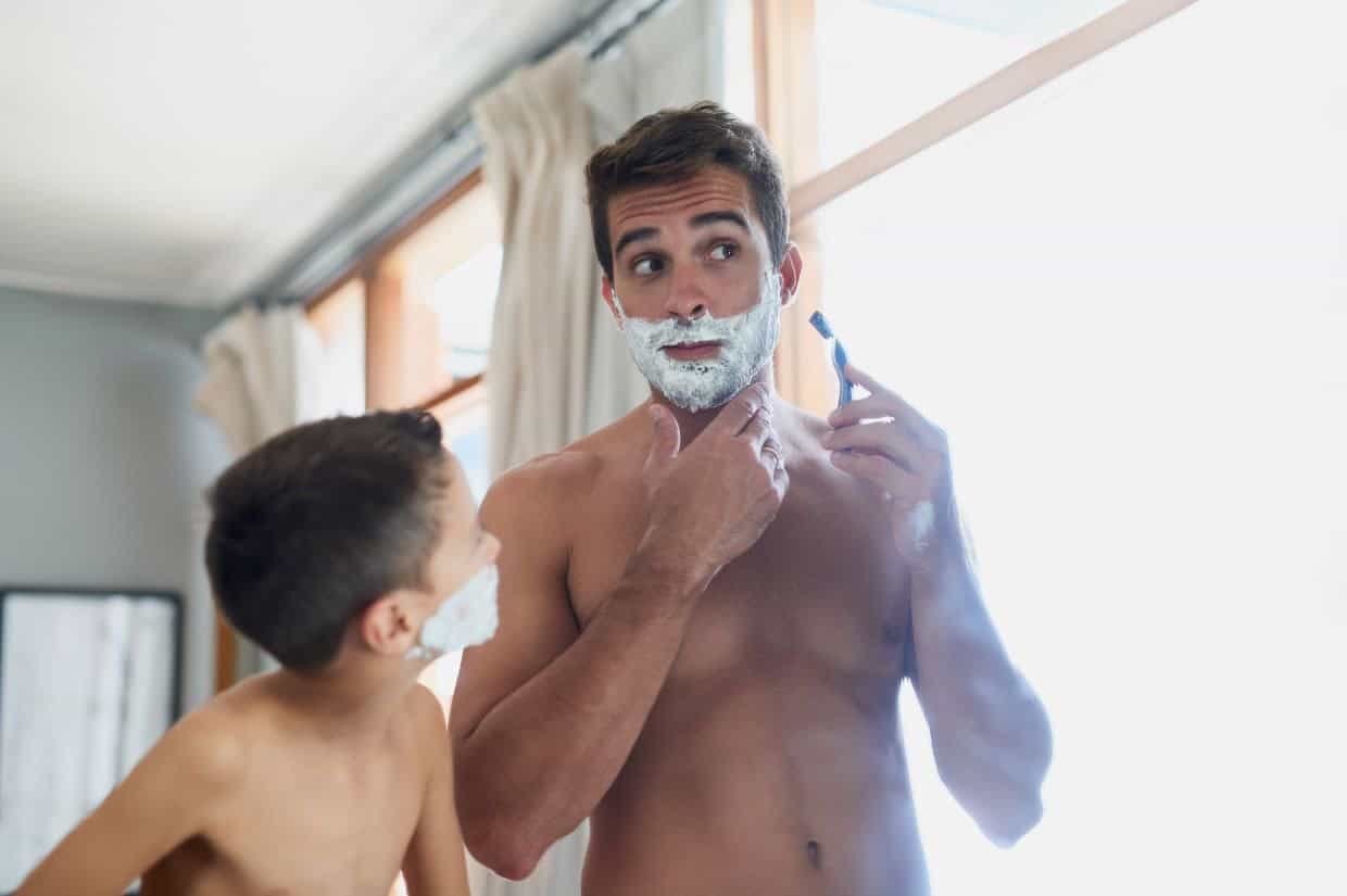 A man and a child apply shaving cream to their faces. The man is holding a razor. They stand in front of a window.