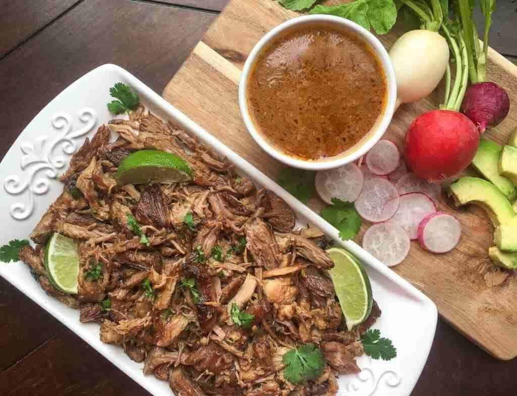 Plate of shredded meat garnished with lime and cilantro, next to sliced radishes and avocado. A bowl of sauce and additional ingredients are on a wooden board.