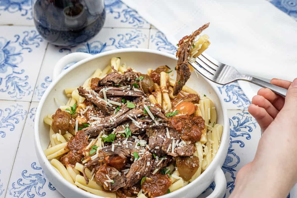 A hand lifting a fork with a bite of Italian Pot Roast Pasta on a bowl.