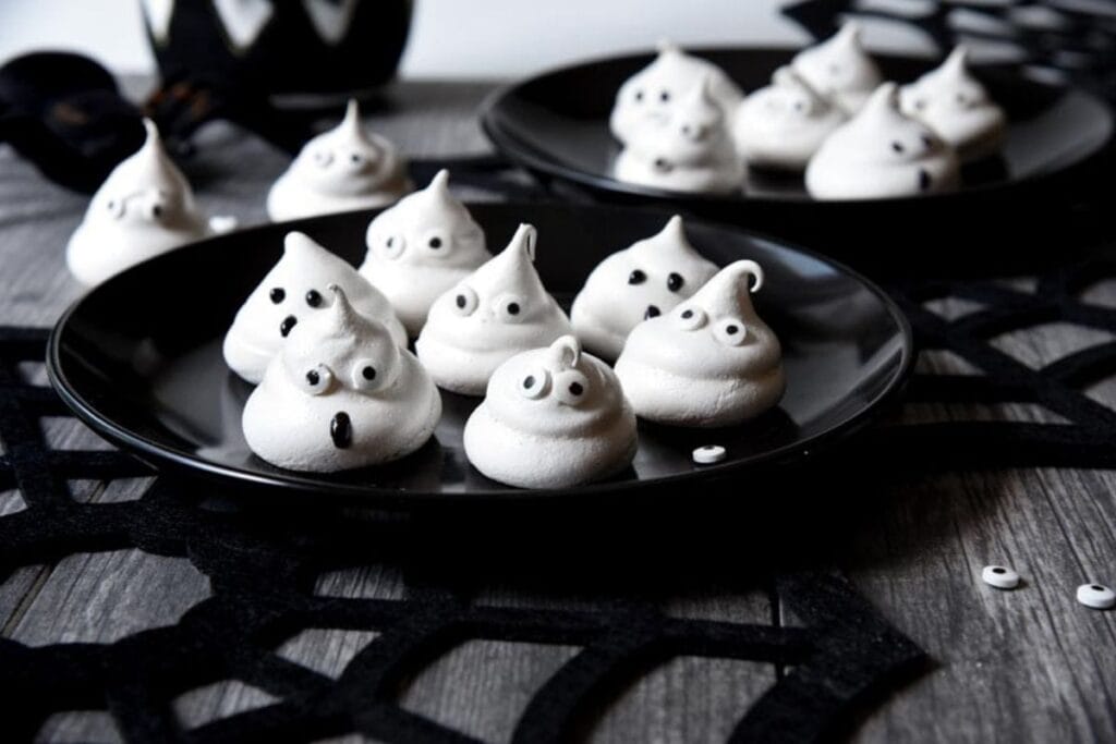 Black plates with ghost-shaped meringues featuring small eyes, set on a spiderweb-themed tablecloth.