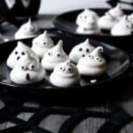 Black plates with ghost-shaped meringues featuring small eyes, set on a spiderweb-themed tablecloth.