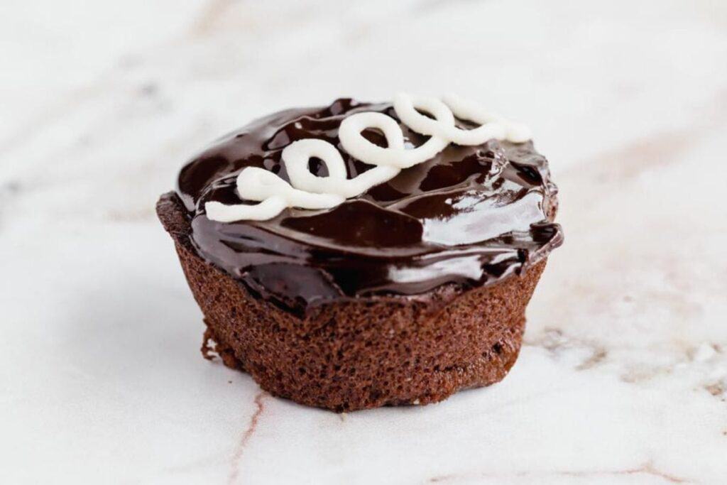 A chocolate cupcake with glossy frosting topped with a white icing swirl, placed on a marble surface.