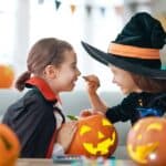 Two children carving a pumpkin. One wears a vampire costume, and the other is dressed as a witch with a black hat.