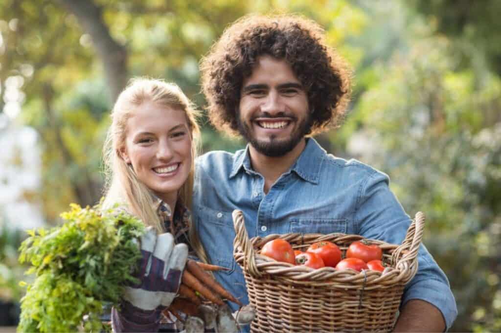 A man and a woman smiling, holding a basket of tomatoes and a bunch of carrots outdoors.