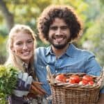 A man and a woman smiling, holding a basket of tomatoes and a bunch of carrots outdoors.