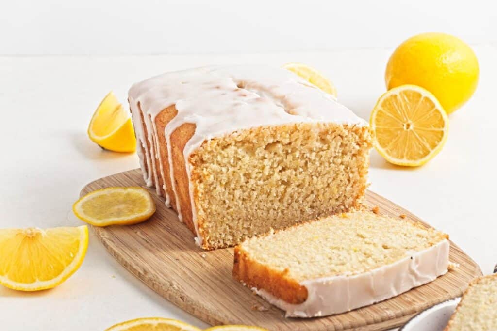 Lemon loaf cake with icing is sliced on a wooden board, surrounded by whole and sliced lemons.